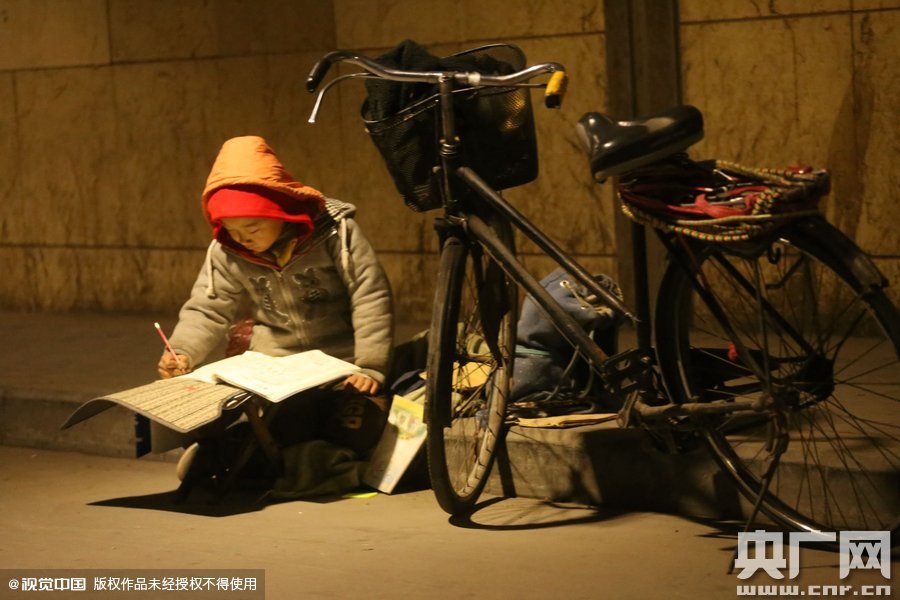 鄭州中原路京廣鐵路大橋下涵洞的馬路牙子邊，一位戴著紅色辣椒帽的小男孩趴在一個小馬扎上，上面鋪著一張車墊，在微弱的燈光下寫作業(yè)