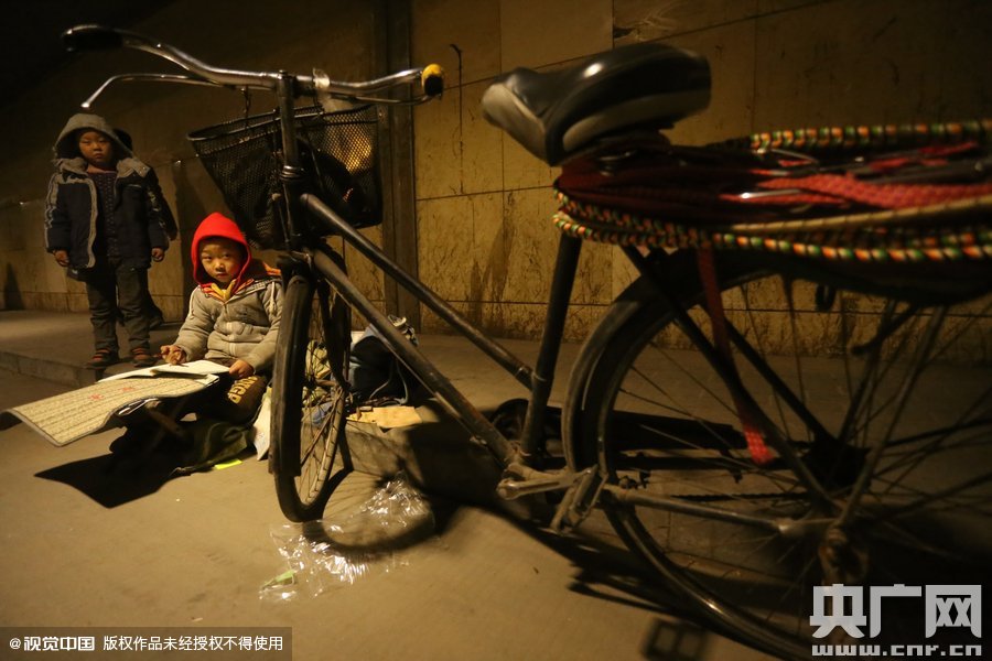 2015年11月8日晚，鄭州，7歲男孩涵洞橋下陪父母練攤，路邊燈下坐小馬扎上寫作業(yè)。